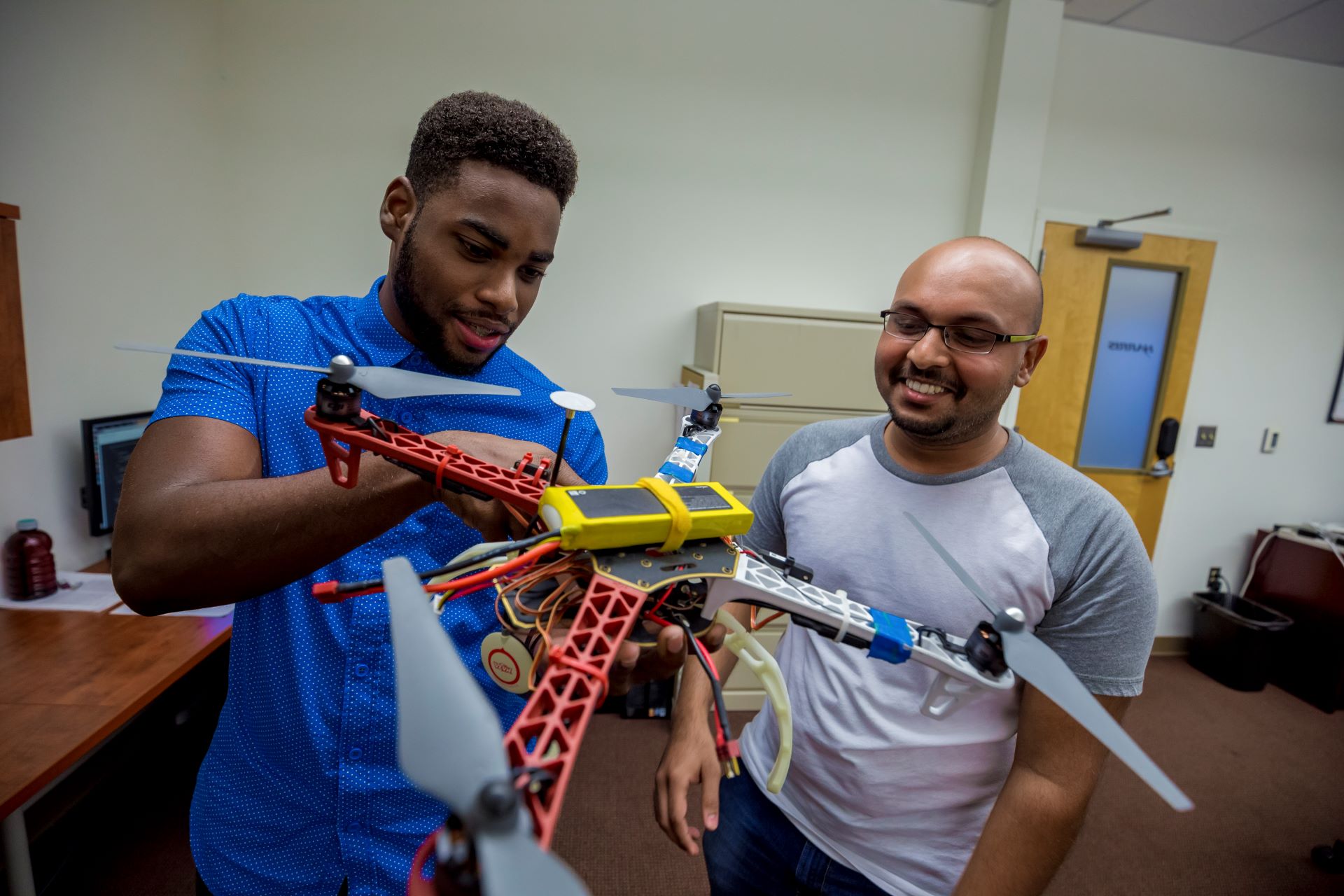 Two students examining a multicolored drone in a classroom, with one student holding it and showing it to the other who is smiling